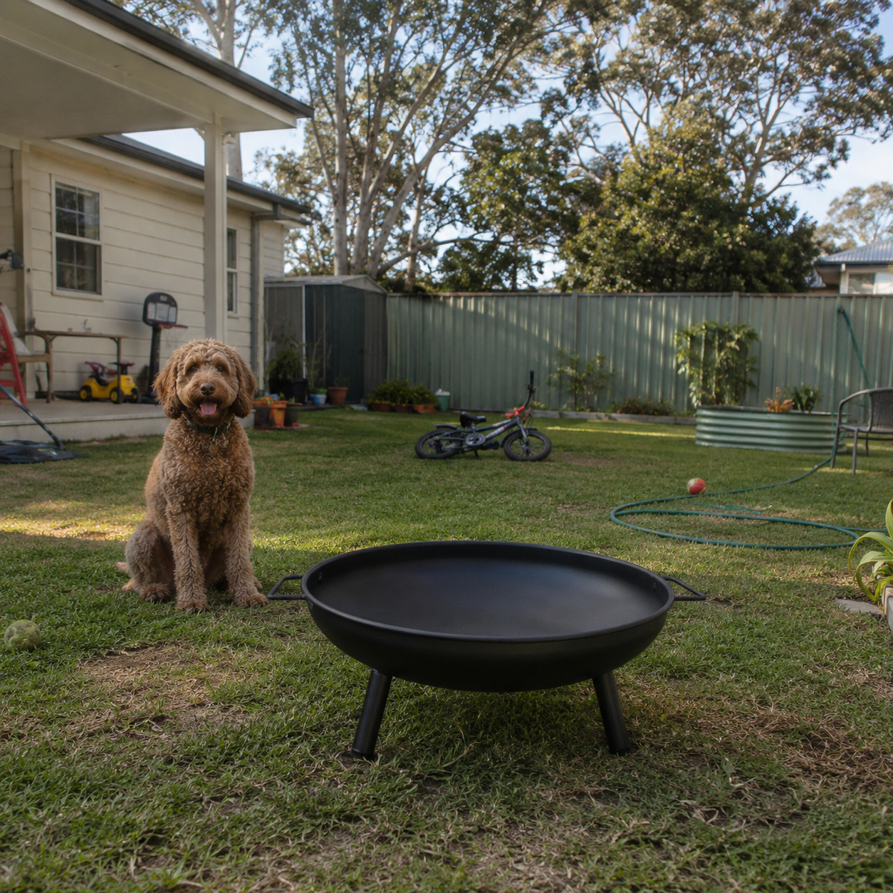 Black steel outdoor fire pit with dog sitting next to it in a backyard with a house and trees in the background.