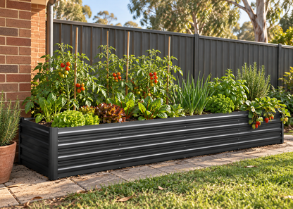 A long black corrugated metal raised garden bed filled with leafy greens, herbs, and tomato plants, situated outdoors next to a brick wall and wooden fence.