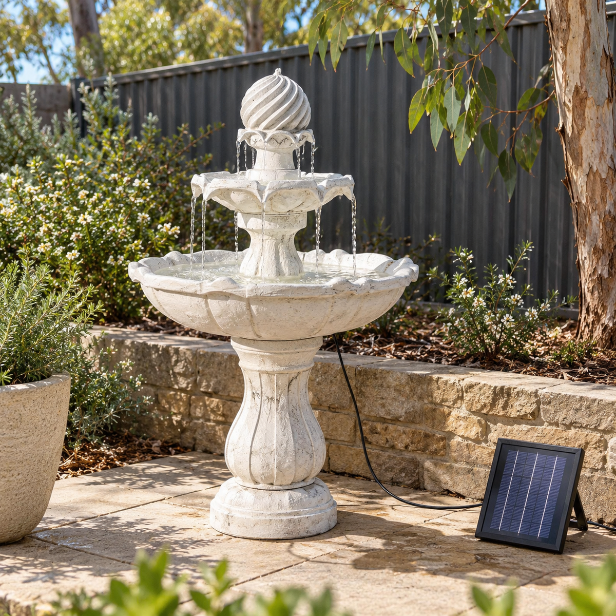 A tiered outdoor stone fountain with water flowing from the top to the basin, placed on a patio beside a small solar-powered charging pad, with shrubs, a fence, and a tree in the background.