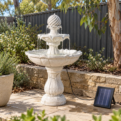 A tiered outdoor stone fountain with water flowing from the top to the basin, placed on a patio beside a small solar-powered charging pad, with shrubs, a fence, and a tree in the background.