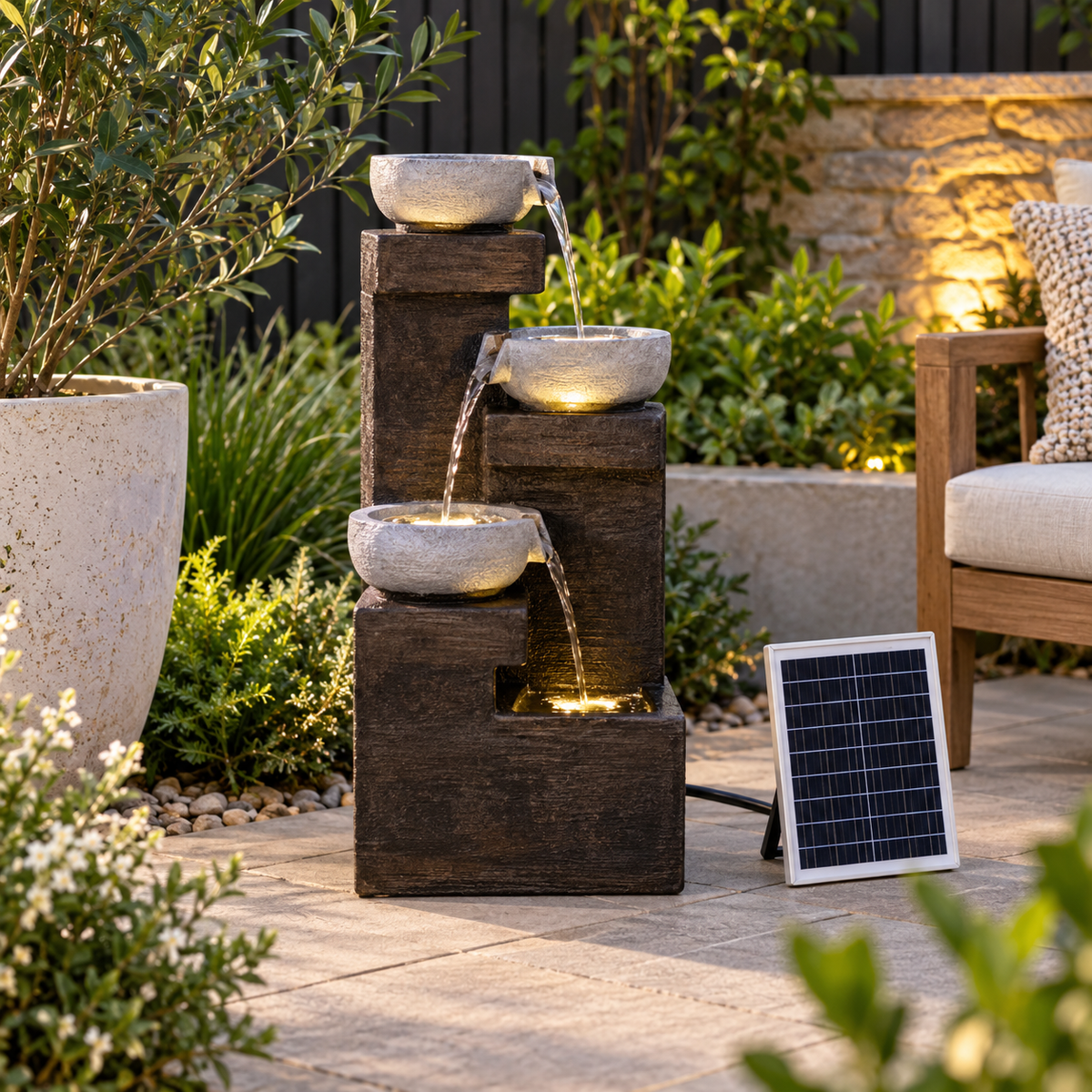 A solar-powered outdoor water fountain with three tiers of dark brown stacked blocks and light gray stone bowls, shown with a small solar panel on a patio next to plants and a chair.