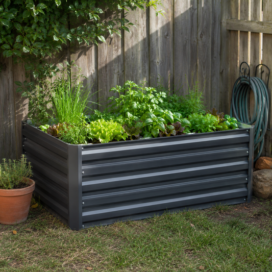 A dark gray corrugated metal raised garden bed filled with green herbs and lettuce, set on a grassy lawn in front of a wooden fence.