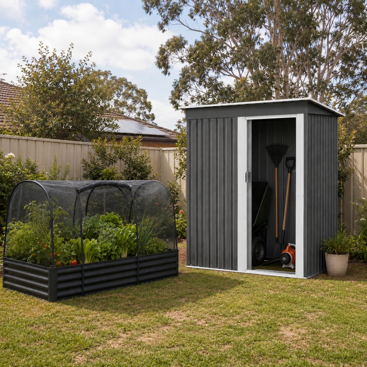 A dark gray metal garden shed with a white-framed door stands on a grassy backyard next to a black raised garden bed with a greenhouse-style mesh cover. A garden rake and other tools are stored inside the shed, with trees and a fence in the background.