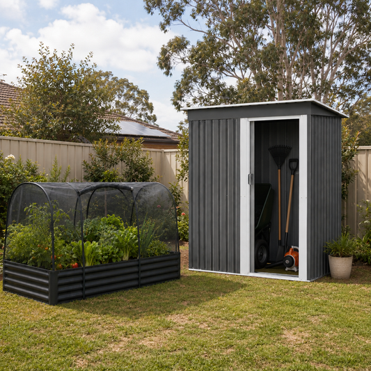 A dark gray metal garden shed with a white-framed door stands on a grassy backyard next to a black raised garden bed with a greenhouse-style mesh cover. A garden rake and other tools are stored inside the shed, with trees and a fence in the background.