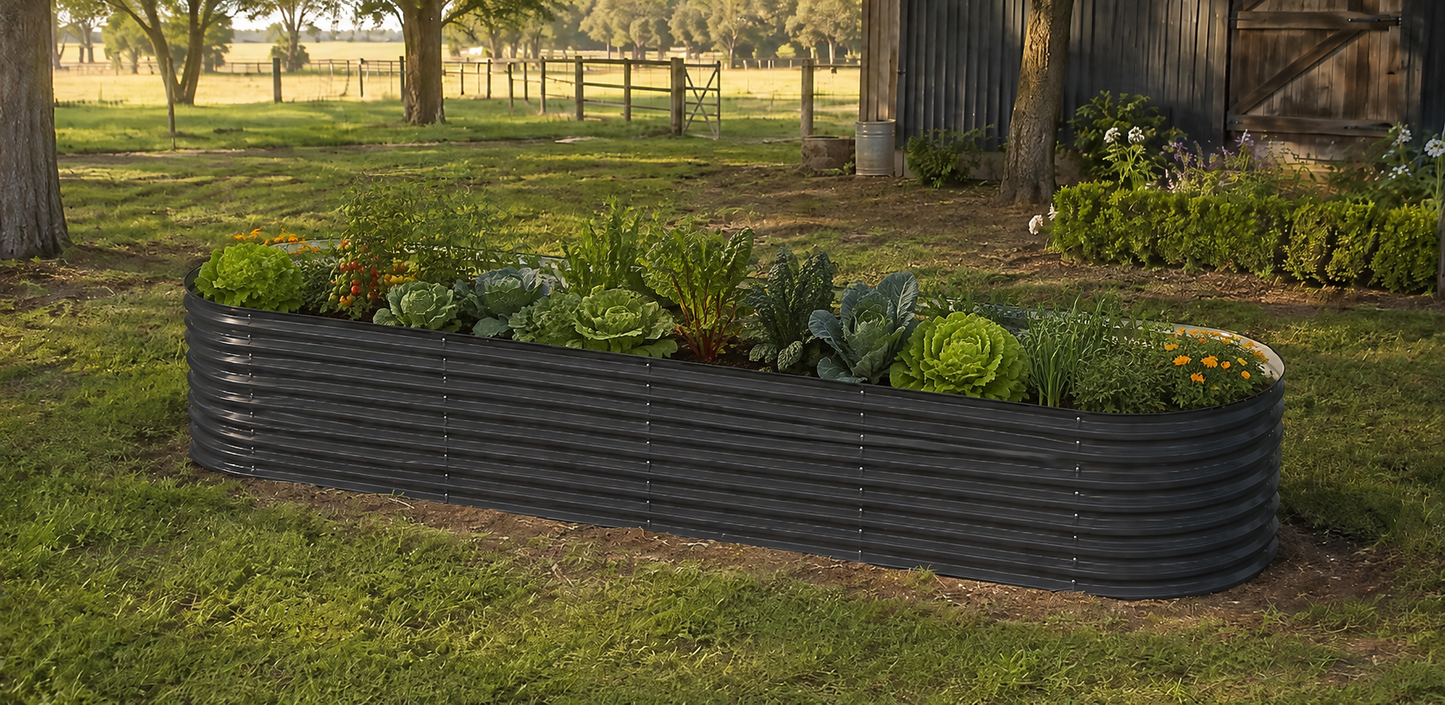 A long, corrugated black raised garden bed filled with green plants and vegetables, situated on a grassy lawn in front of a rustic wooden barn.