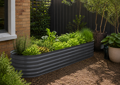 A long, corrugated metal raised garden bed filled with green plants, placed outdoors on a gravel path beside a brick wall and potted trees.