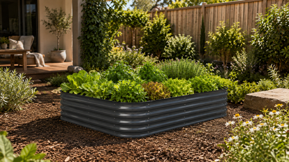 A rectangular raised garden bed made of dark gray corrugated metal, filled with green leafy plants, set in a landscaped backyard garden bed with mulch, shrubs, and a wooden fence in the background.