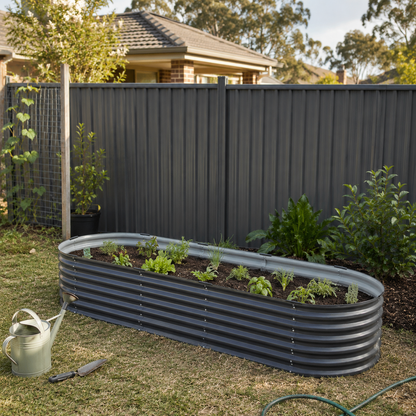 An oval corrugated metal raised garden bed filled with young plants, set in a backyard on grass in front of a dark gray fence. A light green watering can and a hand tool sit nearby on the lawn.