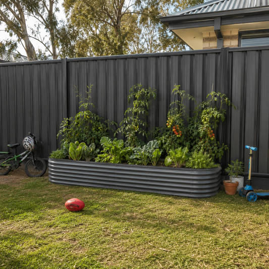 A large corrugated metal raised garden bed filled with leafy greens and tomato plants in a backyard, with a dark gray fence, grass, a bicycle, a red ball, and a scooter nearby.