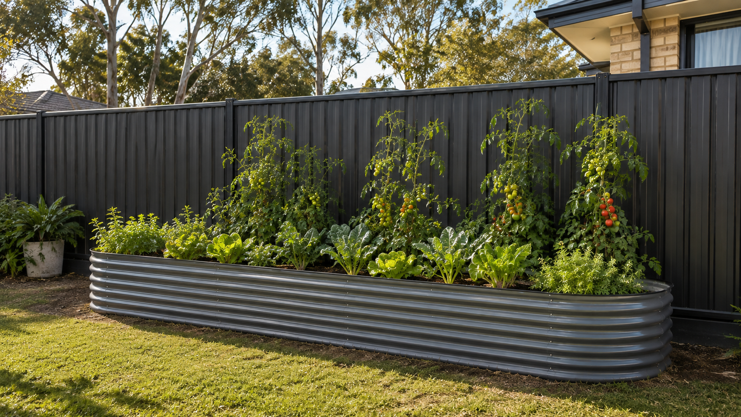 A long corrugated metal raised garden bed filled with leafy greens and tomato plants, set in a backyard with a dark fence, grass, and trees.