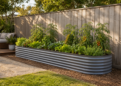 A long corrugated metal raised garden bed filled with leafy greens, tomato plants, and other garden plants, set against a wooden fence in a backyard garden.