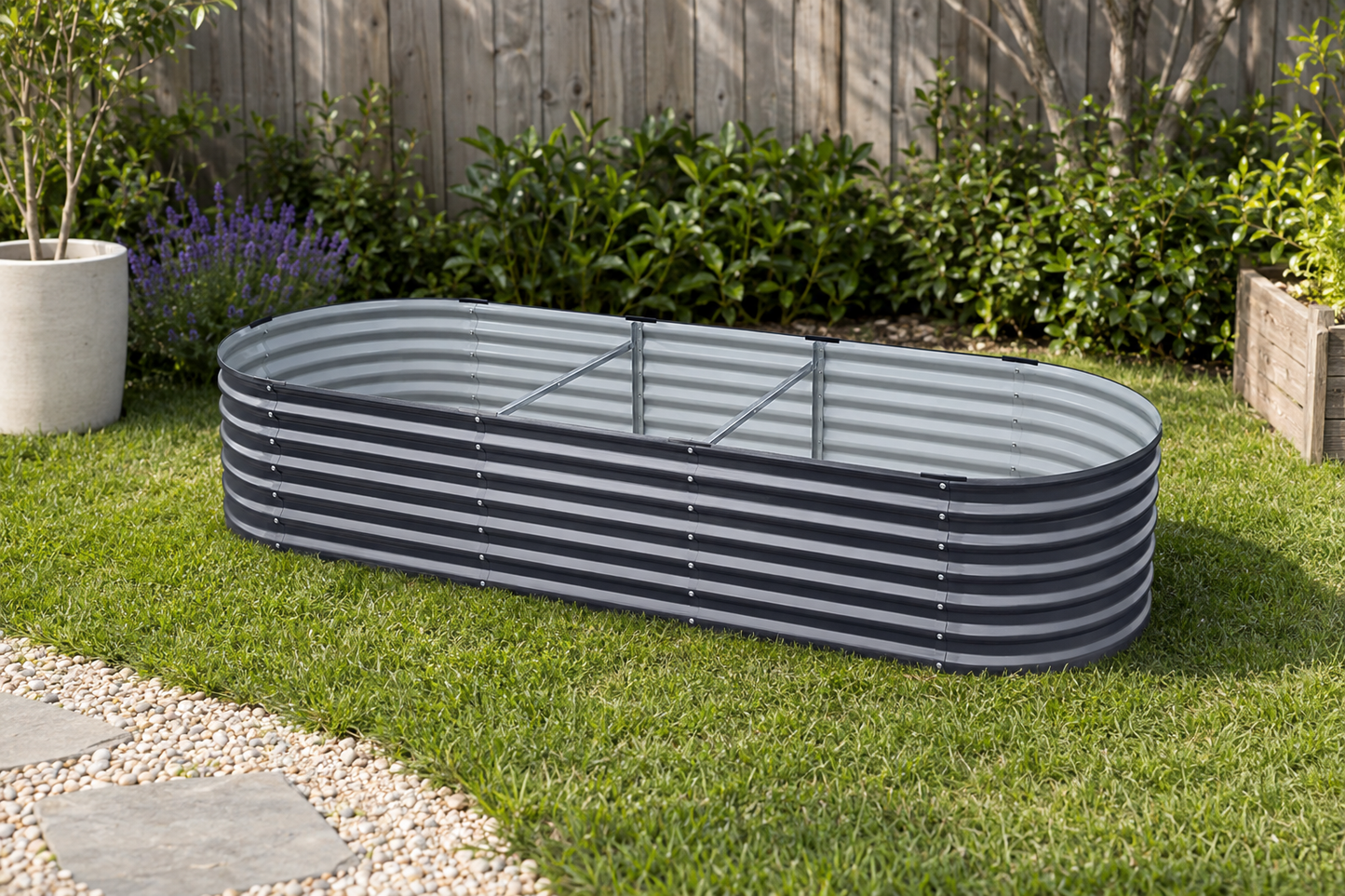 An oval corrugated metal raised garden bed on a green lawn in a backyard setting with a wooden fence, plants, and a stone path.