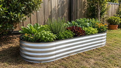 A corrugated galvanized steel raised garden bed filled with various green and red leafy plants, situated in a backyard on grass with a wooden fence in the background.