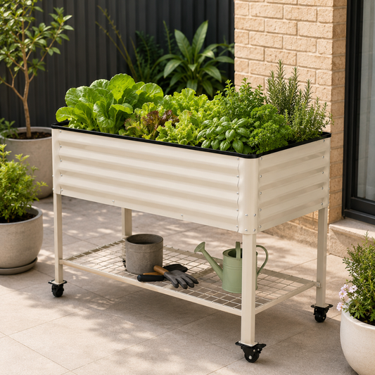 A white corrugated metal raised garden bed on wheels, filled with leafy greens and herbs, set on a patio next to a brick wall with potted plants and gardening tools underneath.