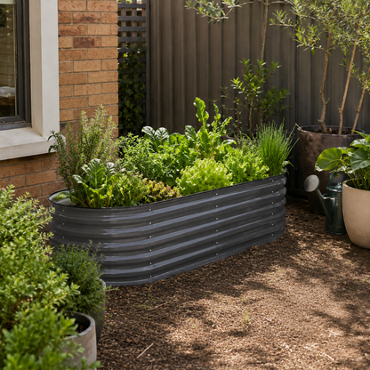 A dark gray corrugated metal raised garden bed filled with green leafy plants, situated in a backyard garden next to a brick wall and mulch ground.