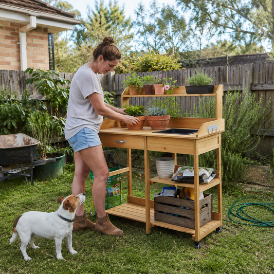 Woman tending to plants in a garden with a wooden potting table and a dog nearby.