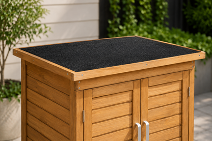 A wooden outdoor storage cabinet with a black textured non-slip top, shown in a garden setting with green plants and a light-colored wall in the background.