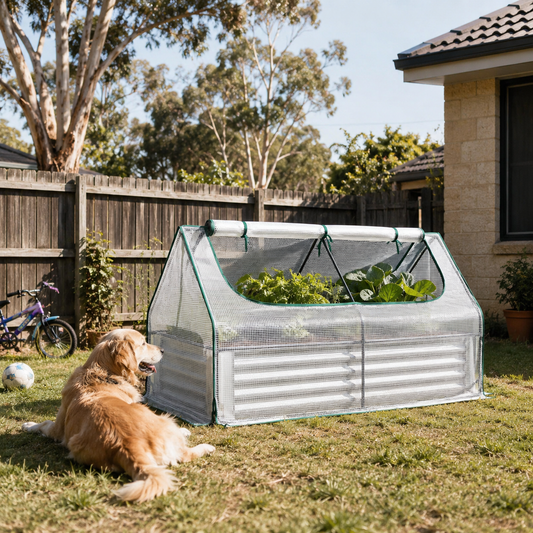 A small pop-up greenhouse with corrugated metal sides and mesh top, set in a backyard on grass next to a golden retriever dog. The greenhouse contains leafy green plants, with a wooden fence, trees, and part of a house visible in the background.