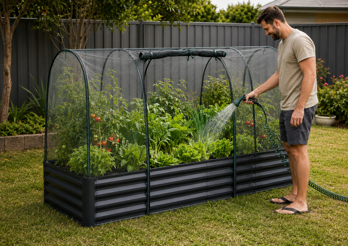 A man watering a raised garden bed covered by a clear arched mesh tunnel in a backyard garden setting.