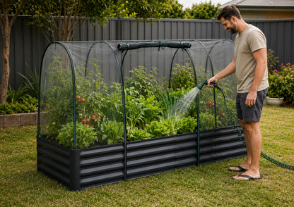 A man watering a raised garden bed covered by a clear arched mesh tunnel in a backyard garden setting.