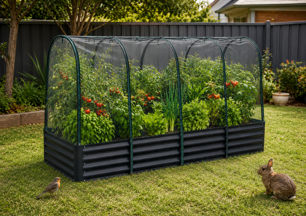 A black corrugated metal raised garden bed with a semi-transparent arched mesh tunnel cover, filled with green plants and red flowers, set in a grassy backyard with a bird and rabbit nearby.