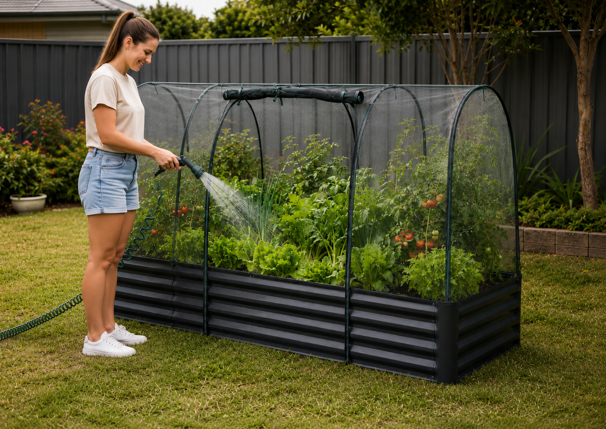 A woman watering a raised garden bed with a mini hoop house in a backyard garden setting.