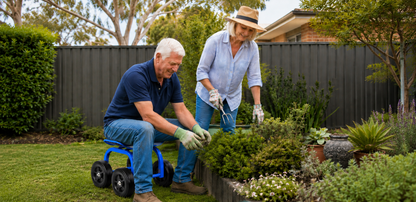 An older man and woman gardening in a backyard. The man is kneeling on a blue wheeled lawn chair, wearing a navy polo shirt, blue jeans, and green gloves while trimming a green bush.