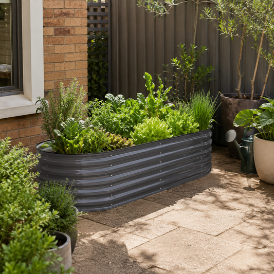 A dark gray corrugated metal raised garden bed filled with green herbs and plants, situated on a stone patio next to a brick house and other potted plants.
