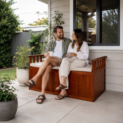 A man and a woman sit on a wooden outdoor storage bench with a cushioned seat, holding mugs on a patio in front of a house. The bench has a warm brown wood finish and a light beige cushion.