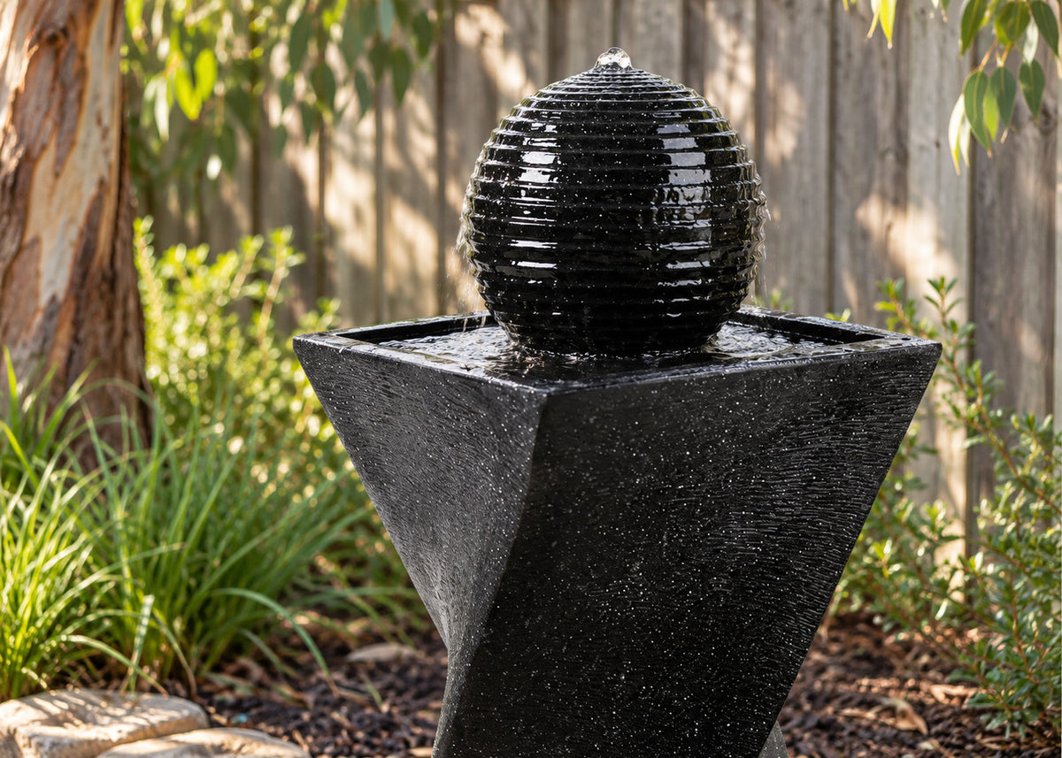 A black spherical water fountain with a ribbed, textured surface sits on a matching geometric black pedestal in a garden setting with plants and a wooden fence in the background.