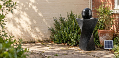 A black stone fountain sculpture with a spherical top on a twisted pedestal, set outdoors on a stone patio beside green shrubs, a potted plant, and a small solar panel against a cream brick wall.