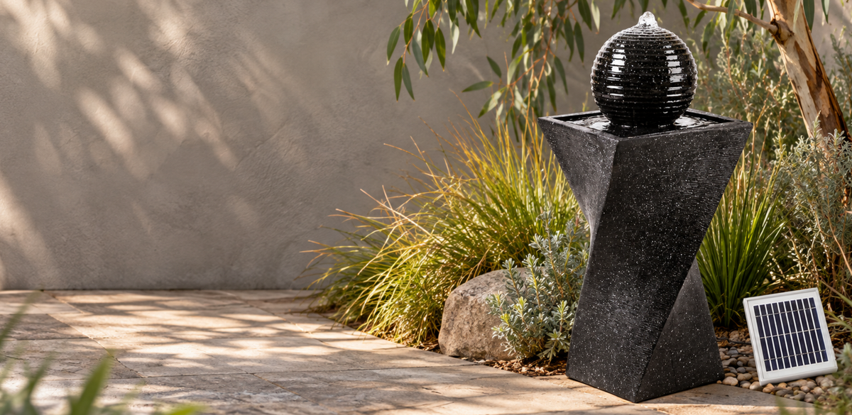 A black solar-powered fountain with a textured spherical top and geometric pedestal base, set outdoors in a garden next to a small solar panel.