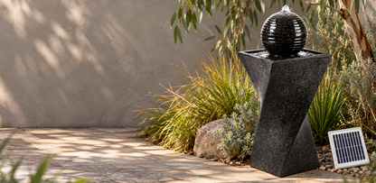 A black solar-powered fountain with a textured spherical top and geometric pedestal base, set outdoors in a garden next to a small solar panel.