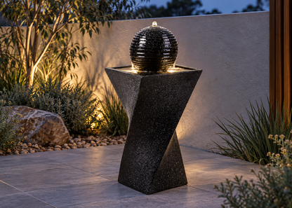 A modern black stone fountain with a ribbed spherical top and geometric pedestal base, set outdoors on a tiled patio at dusk with warm lighting and plants in the background.