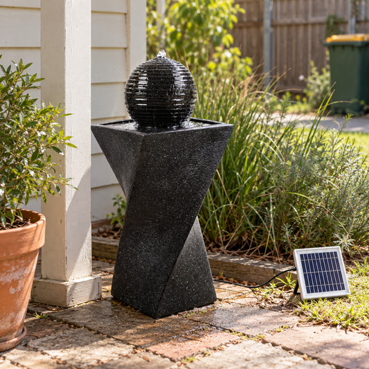 A black solar-powered outdoor fountain with a textured spherical top and geometric pedestal base, placed on a brick patio next to a potted plant in a garden setting.