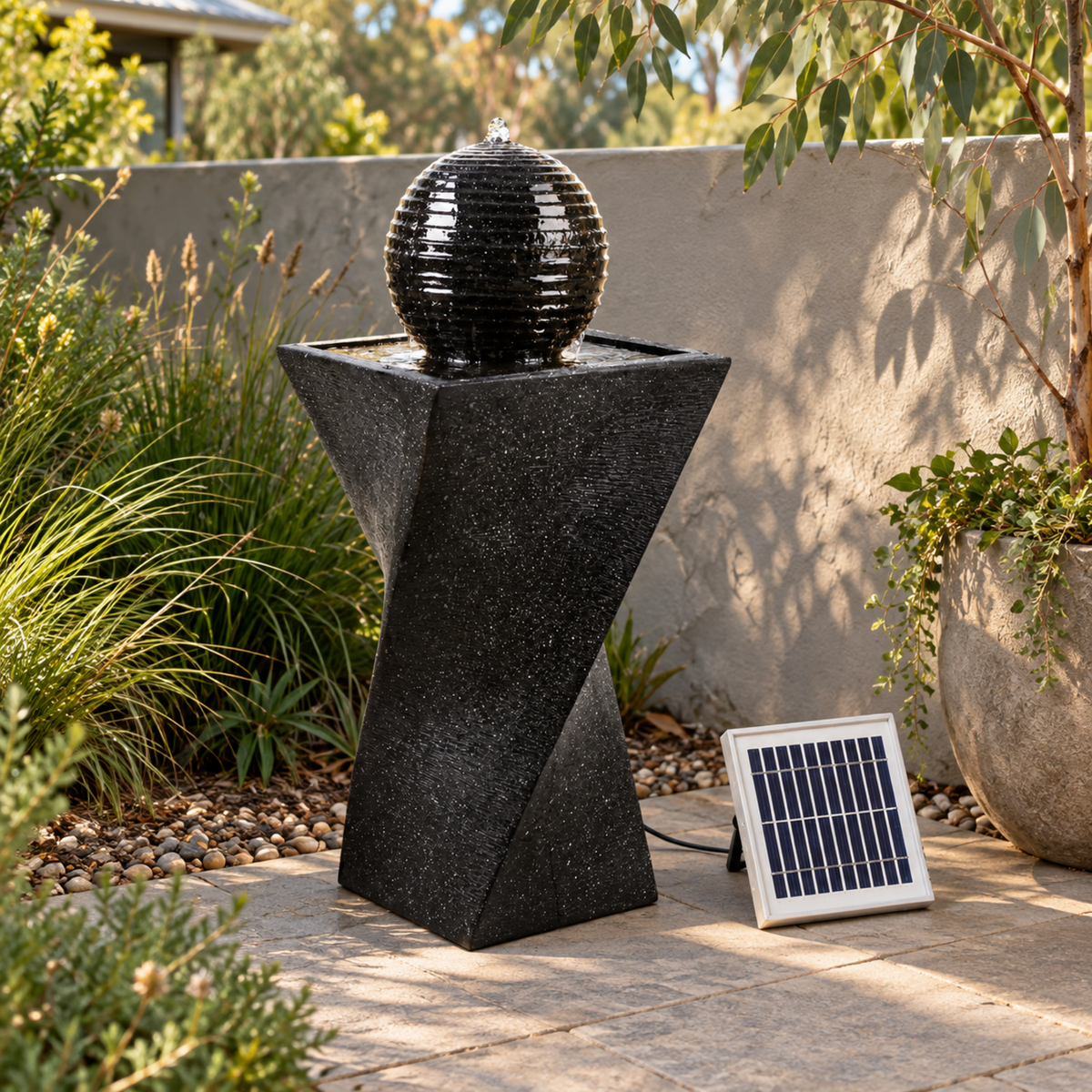 A black solar-powered outdoor water fountain with a textured spherical pool on a geometric pedestal, shown next to its solar panel in a landscaped garden setting.
