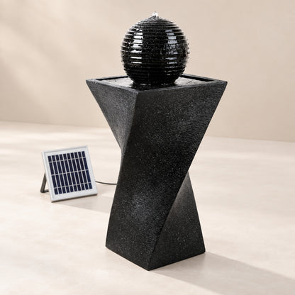 A black spiral pedestal fountain with a spherical water feature on top, shown with a small solar panel on a beige studio background.