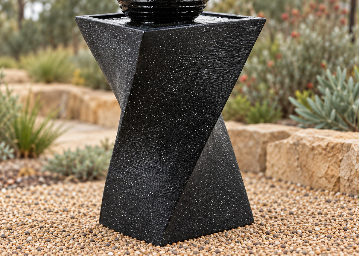 A black textured stone fountain base with a geometric spiral design, shown outdoors on a bed of small tan gravel with a blurred garden background.