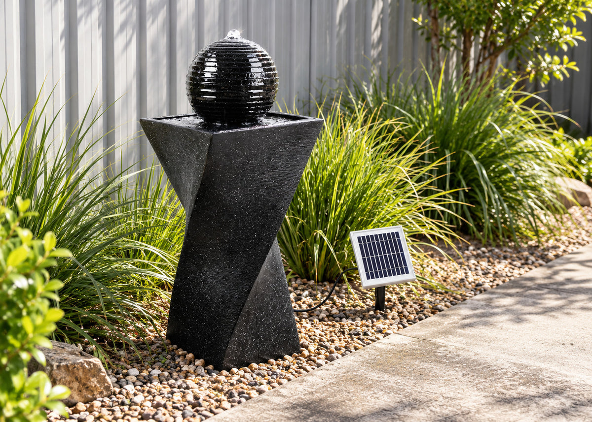 A black geometric stone fountain with a spherical water feature on top, set in a landscaped garden bed beside a concrete path with a small solar panel nearby.