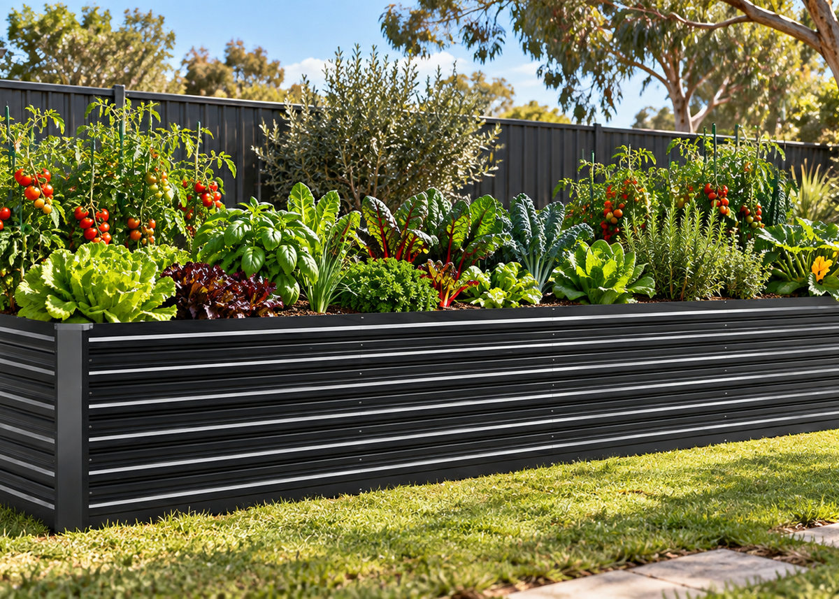 A grey corrugated metal raised garden bed filled with green lettuce, kale, herbs, and red tomato plants, set in a backyard with a grassy lawn, fence, and trees.