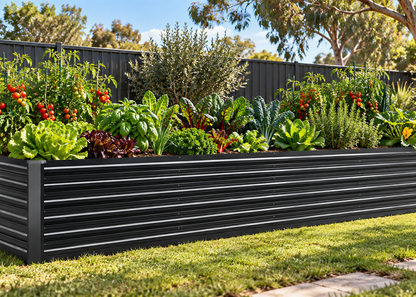A grey corrugated metal raised garden bed filled with green lettuce, kale, herbs, and red tomato plants, set in a backyard with a grassy lawn, fence, and trees.
