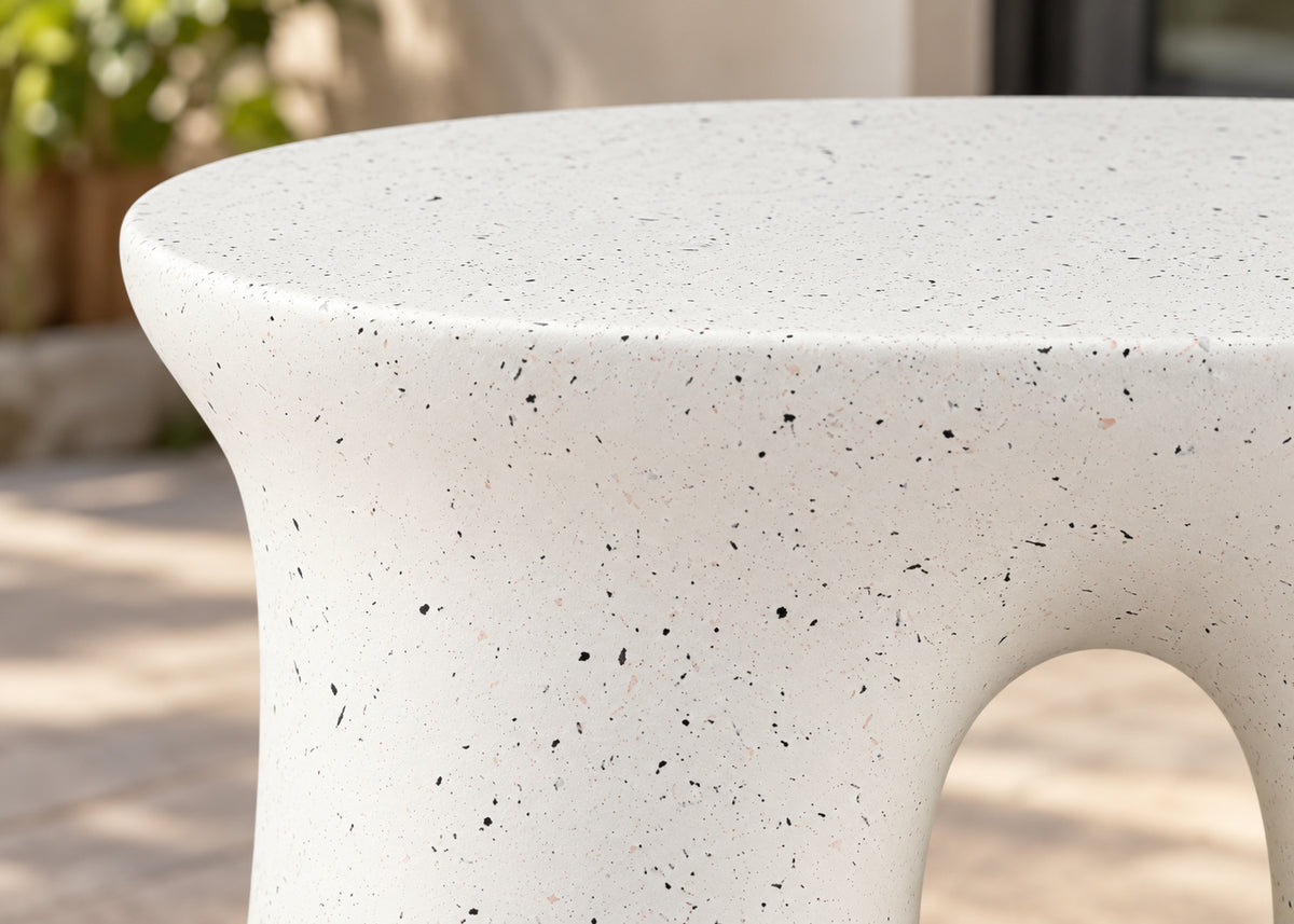 Close-up of a round outdoor stone table with a textured off-white surface and small dark speckles, showing a curved base and a cutout opening.