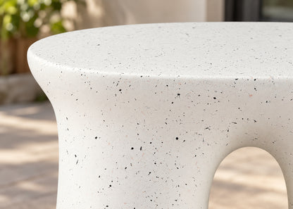 Close-up of a round outdoor stone table with a textured off-white surface and small dark speckles, showing a curved base and a cutout opening.