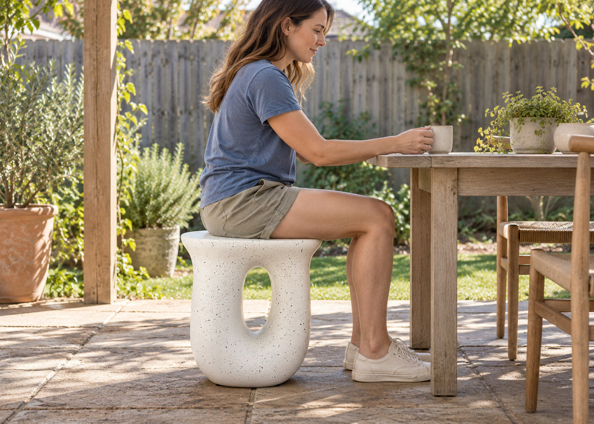 A woman sits on a round concrete stool with a cutout center, placed beside a wooden outdoor dining table on a patio.
