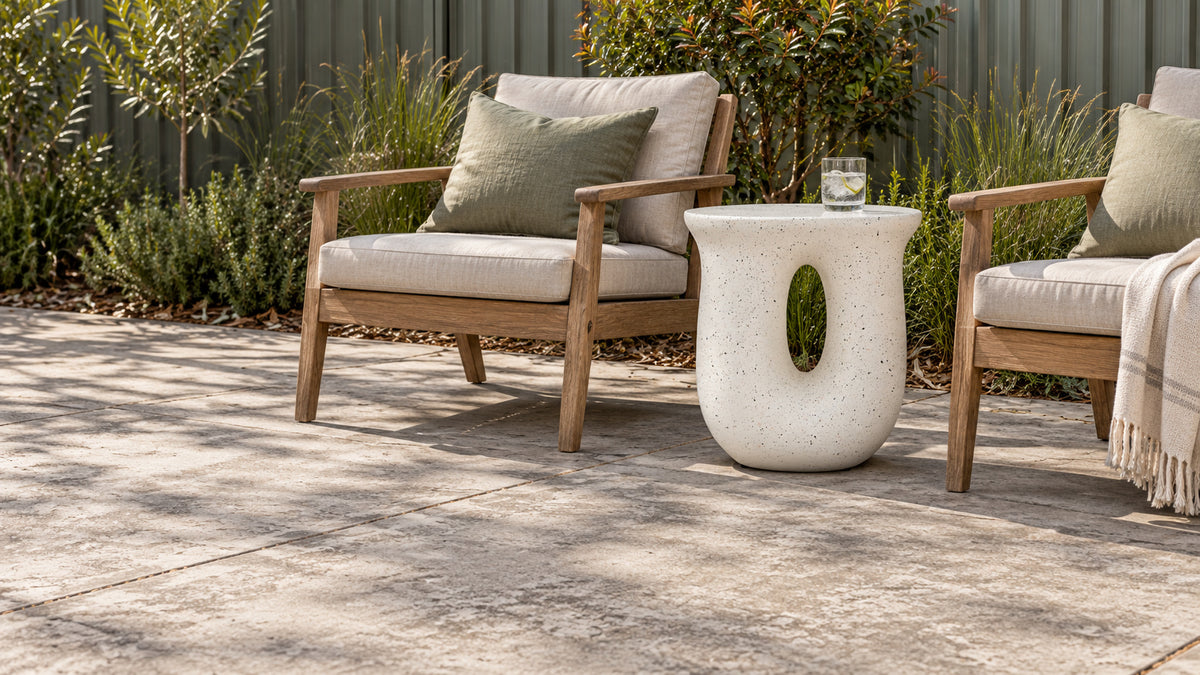 Outdoor patio furniture set featuring two wooden lounge chairs with cream cushions and a matching round concrete side table, styled on a stone patio with greenery in the background.
