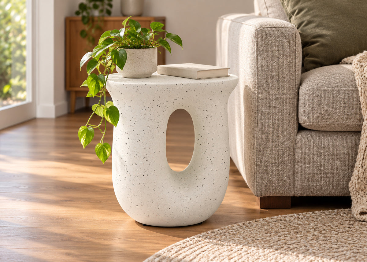 A modern off-white speckled ceramic side table with a large oval cutout, styled in a living room next to a beige sofa with a trailing green plant on top.