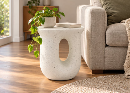 A modern off-white speckled ceramic side table with a large oval cutout, styled in a living room next to a beige sofa with a trailing green plant on top.