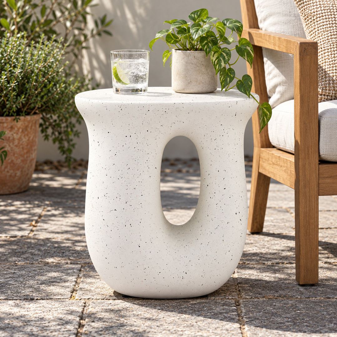 A white speckled concrete side table with an oval cutout, styled outdoors on a stone patio next to a wooden chair with a potted plant on top.