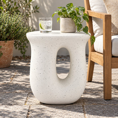 A white speckled concrete side table with an oval cutout, styled outdoors on a stone patio next to a wooden chair with a potted plant on top.