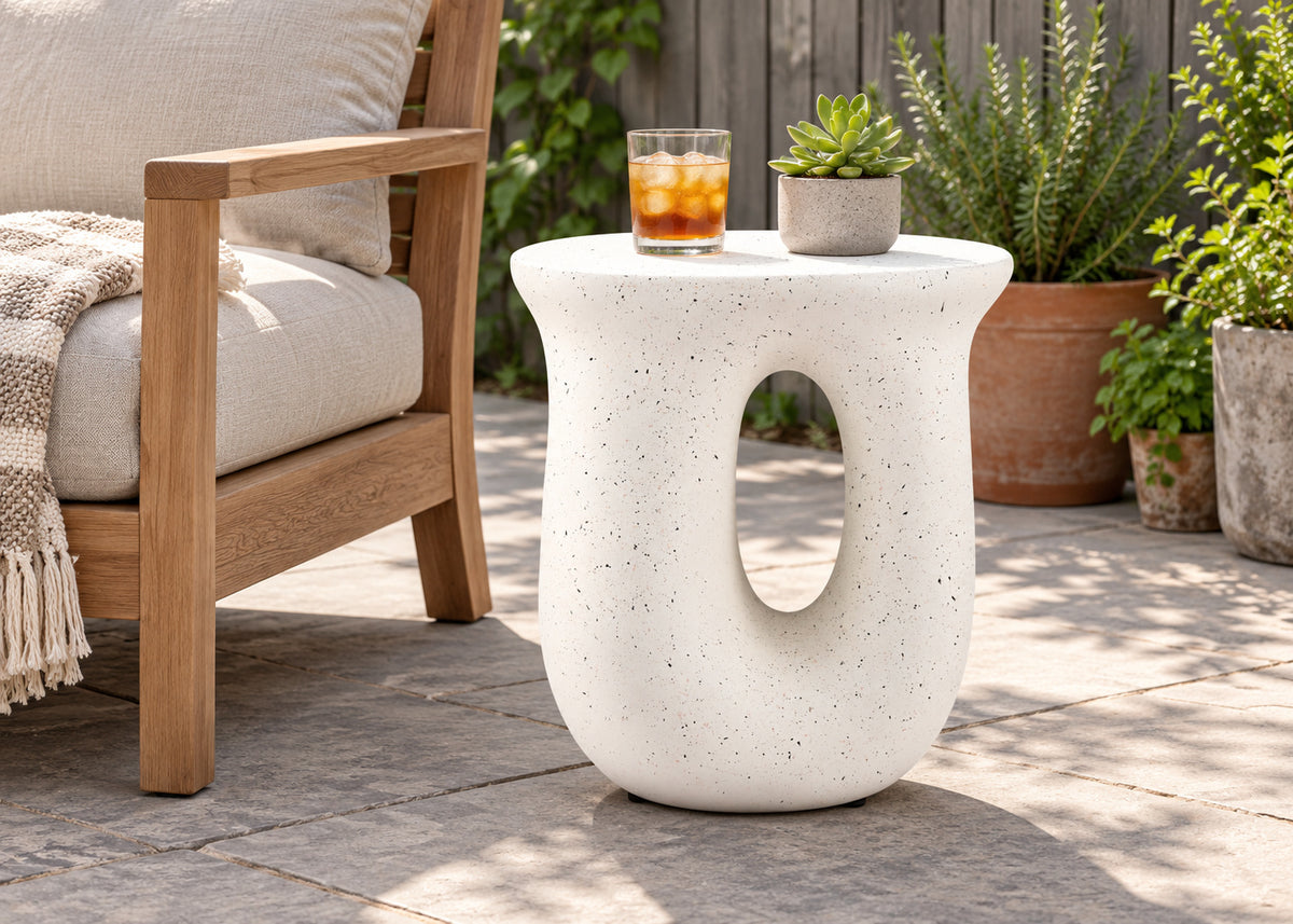 A modern outdoor side table with a rounded, sculptural shape and a central cutout, made from speckled off-white stone. It is styled on a stone patio next to a wooden armchair with a throw blanket, with potted green plants in the background.